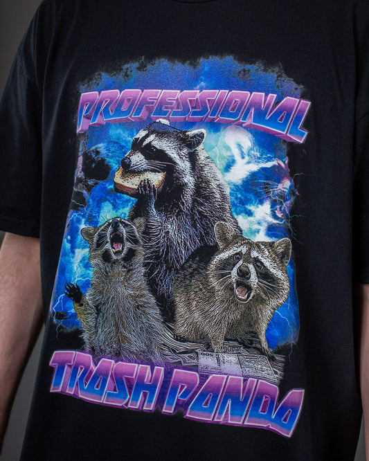 Frontalansicht eines schwarzen T-Shirts mit auffälligem Waschbär-Motiv. Die tragende Person zieht den unteren Saum leicht zur Seite. Über den drei Waschbären steht „PROFESSIONAL“, darunter „TRASH PANDA“, gestaltet im leuchtenden Farbverlauf von Pink bis Blau.