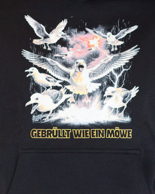Detailansicht des Frontprints auf einem schwarzen Kapuzenpullover: Mehrere wütend wirkende Möwen mit ausgebreiteten Flügeln in einer stürmischen Szenerie. Unten der Schriftzug „GEBRÜLLT WIE EIN MÖWE“ in fetten, gelben Großbuchstaben.