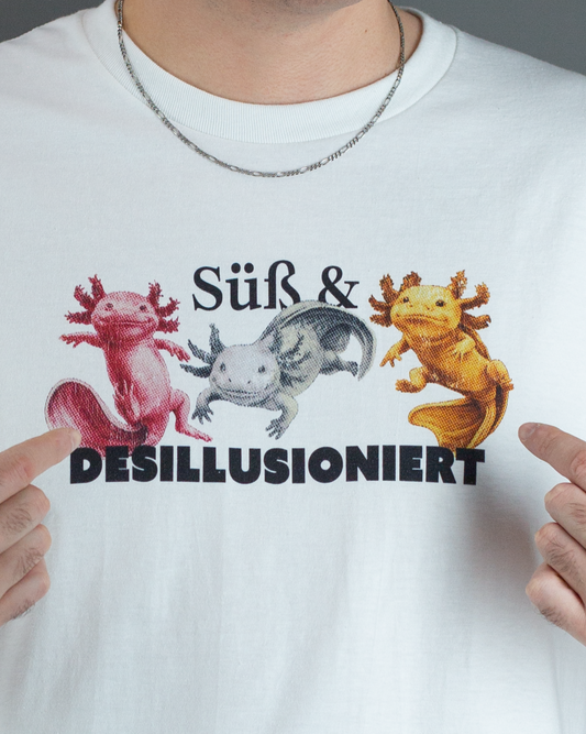 Nahaufnahme des Druckmotivs auf einem weißen T-Shirt mit drei farbigen Axolotl (Rot, Grau, Gelb) und dem zweizeiligen Schriftzug „Süß & DESILLUSIONIERT“.