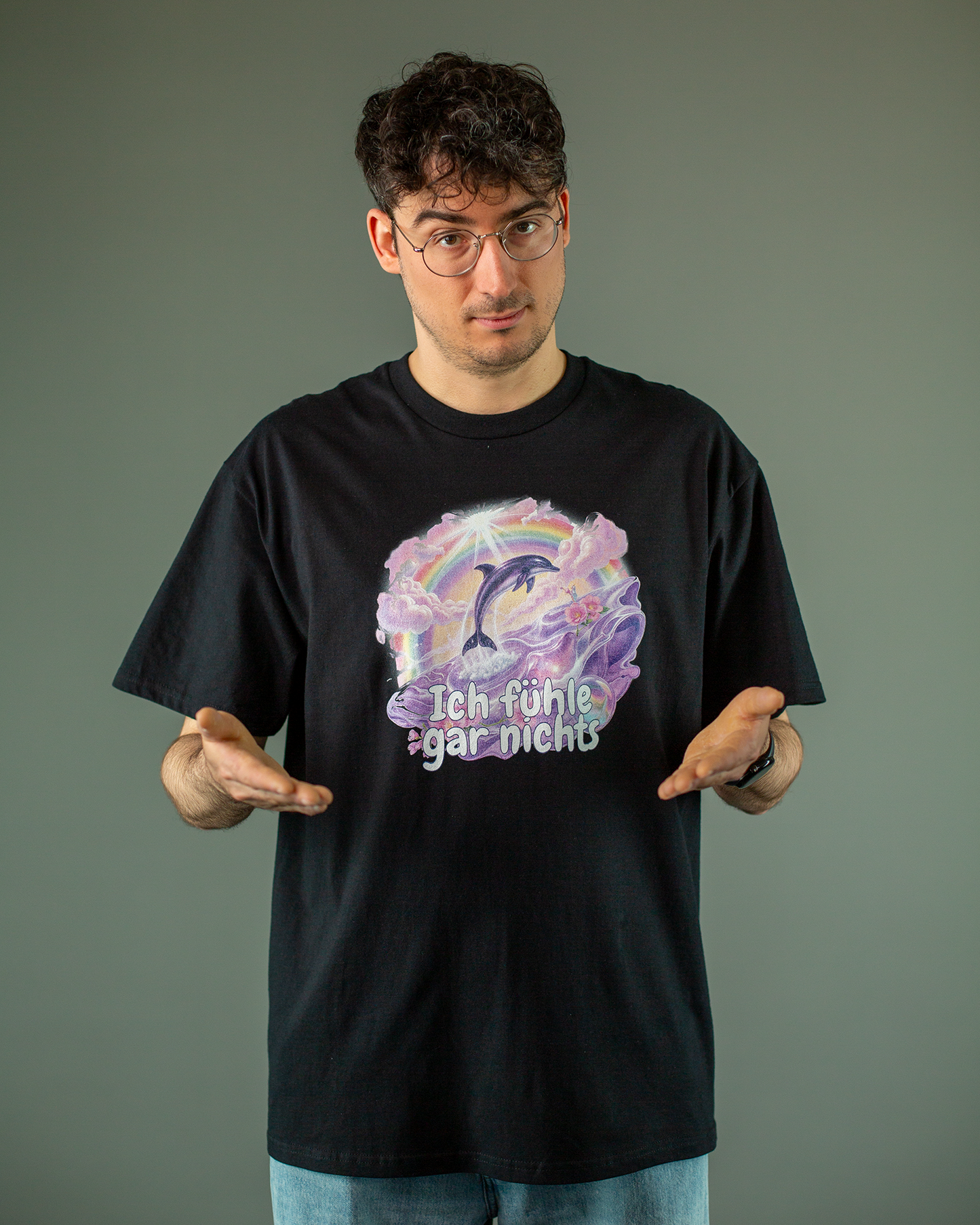 Schwarzes T-Shirt mit großem Frontprint in Pastellfarben. Das Design zeigt einen springenden Delfin vor einem Regenbogen und rosa Wolken. Im Vordergrund schwimmen lila Meerestiere in einer stilisierten Wasserlandschaft. Darunter steht in verspielter Schrift der Text „Ich fühle gar nichts“