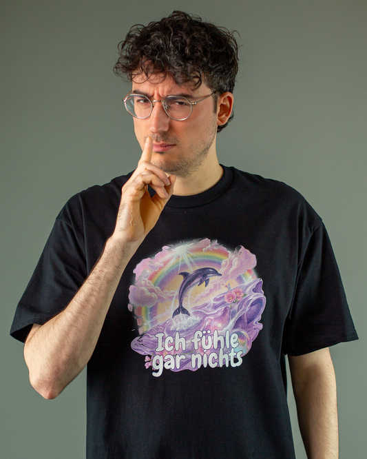 Schwarzes T-Shirt mit farbenfrohem Print auf der Vorderseite. Das Motiv zeigt einen springenden Delfin, Regenbogen, Wolken und ein verspieltes Meeresszenario in Lila und Rosa. Unter dem Bild steht der Schriftzug „Ich fühle gar nichts“ in großer, kindlich wirkender Schrift.