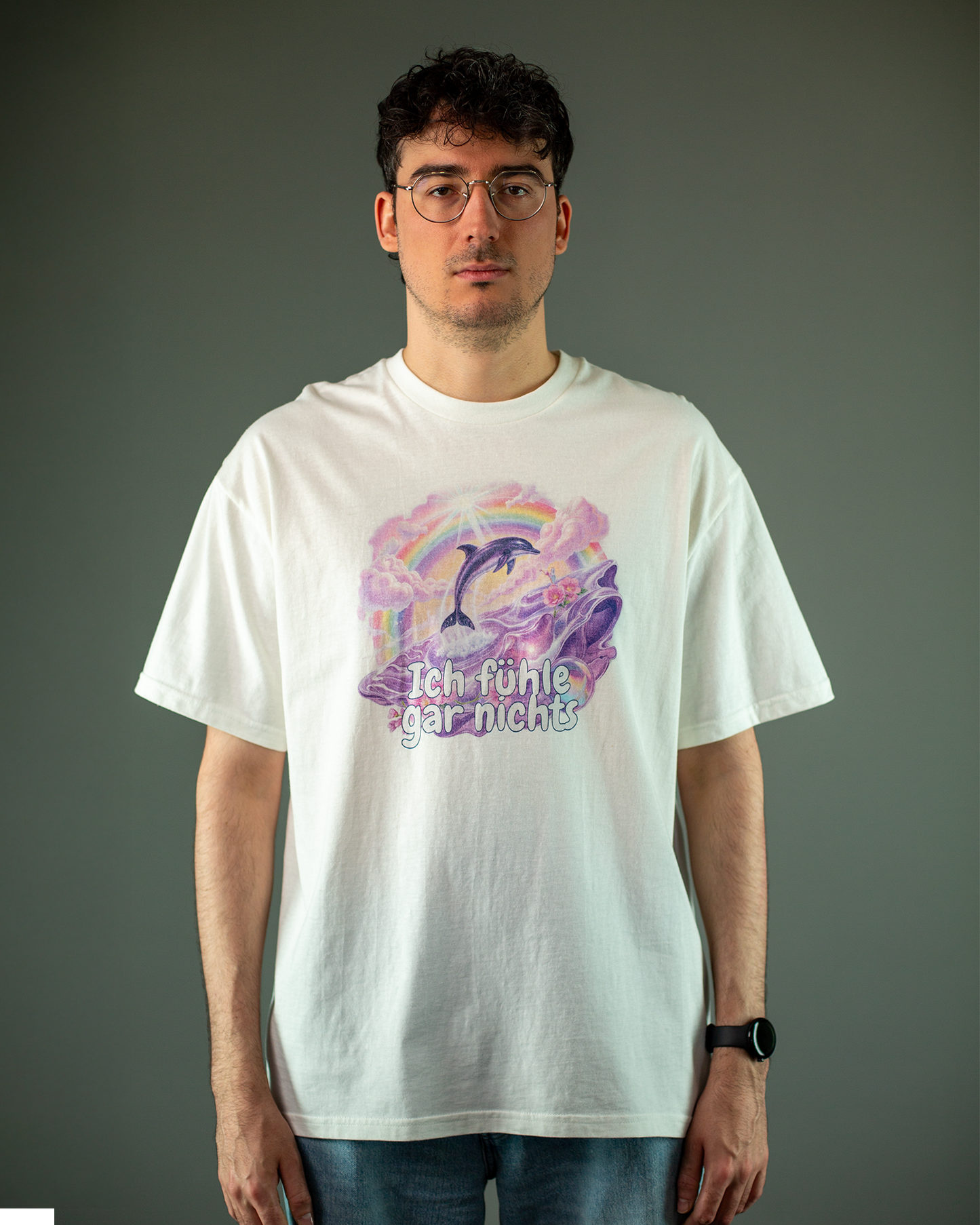 Weißes T-Shirt mit großem Frontprint in Pastellfarben. Das Design zeigt einen springenden Delfin vor einem Regenbogen und rosa Wolken. Im Vordergrund schwimmen lila Meerestiere in einer stilisierten Wasserlandschaft. Darunter steht in verspielter Schrift der Text „Ich fühle gar nichts“