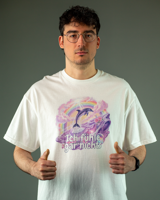 Weißes T-Shirt mit farbenfrohem Print auf der Vorderseite. Das Motiv zeigt einen springenden Delfin, Regenbogen, Wolken und ein verspieltes Meeresszenario in Lila und Rosa. Unter dem Bild steht der Schriftzug „Ich fühle gar nichts“ in großer, kindlich wirkender Schrift.