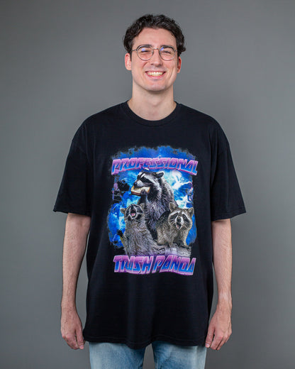 Schwarzes T-Shirt mit farbigem Frontprint, getragen von einer Person frontal zur Kamera. Der Druck zeigt drei Waschbären vor einem dramatisch inszenierten blauen Himmel mit Blitzen. Der mittlere Waschbär hält ein Sandwich. Darüber steht der Schriftzug „PROFESSIONAL“, darunter „TRASH PANDA“ in lila-blauem 3D-Stil.