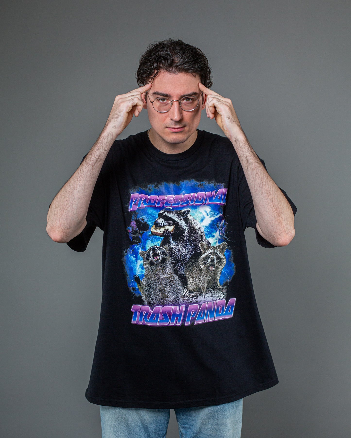 Detailansicht des Frontprints auf schwarzem T-Shirt. Zu sehen sind drei Waschbären, von denen einer ein Sandwich hält, dazu der Schriftzug „PROFESSIONAL TRASH PANDA“ in auffälliger pink-blauer Typografie. Der Hintergrund zeigt ein Blitzgewitter.