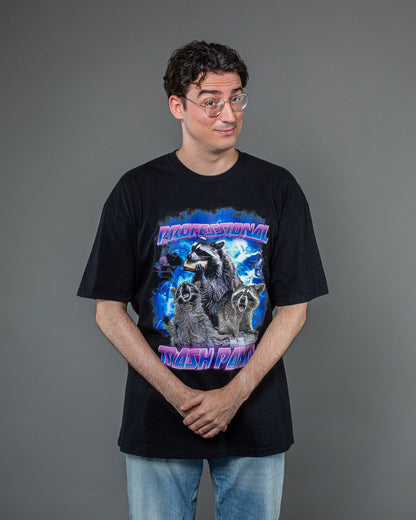Schwarzes T-Shirt mit dreiteiligem Waschbär-Motiv und Schriftzug „PROFESSIONAL TRASH PANDA“, getragen von einer Person, die mit verschränkten Händen leicht schräg zur Kamera steht. Der Druck hebt sich farblich kräftig vom schwarzen Stoff ab.